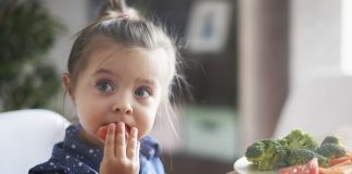 Mitos da alimentação infantil: deixe truques de lado e observe sinais do seu filho; menina de cabelo preso come tomate e tem prato à frente com tomate, brócolis e cenoura