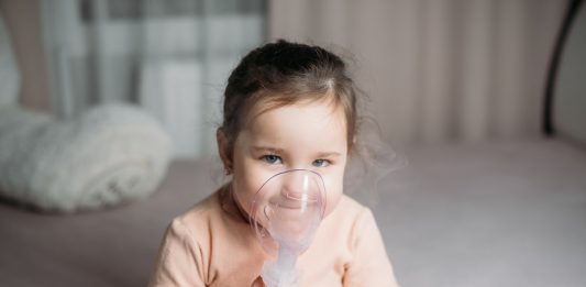 Na foto, criança sentada segurando máscara de nebulização em frente ao rosto enquanto sorri