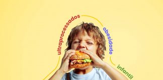 Alimentos ultraprocessados na infância pioram obesidade na vida adulta; menino de camiseta branca e suspensório dá mordida em um sanduíche de hambúrguer