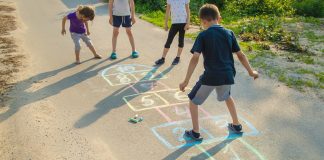 Brincadeiras populares para reviver a infância com os filhos; quatro crianças brincam de amarelinha desenhada no chão da rua com giz de cera