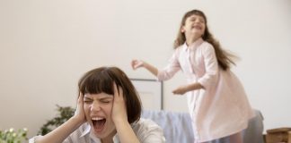 Os gritos dos pais: alguns dos motivos que levam a isso; mãe grita com mãos na cabeça e filha aparece ao fundo rindo