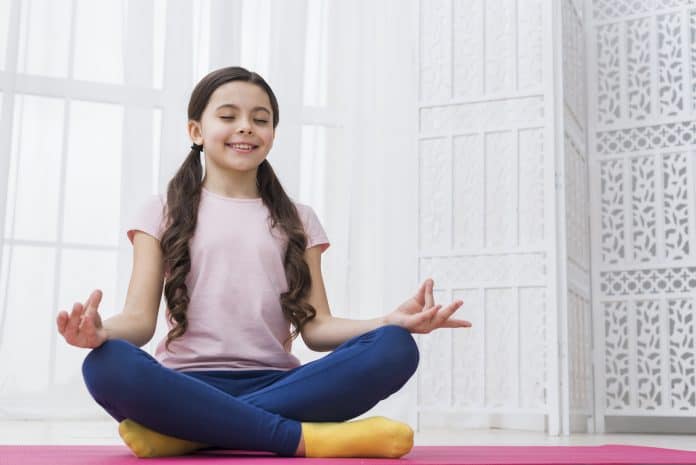 Meditação teleguiada para crianças: saiba onde praticar; menina de cabelo grande dividido ao meio está sentada no chão de olhos fechados e mãos sobre as pernas
