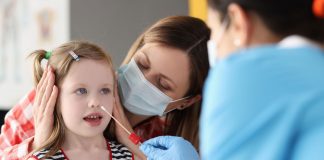 Teste de Covid-19 em criança: o que considerar para realizar o exame; menina sorridente segura teste de Covid-19; mãe acompanha filha que faz teste de Covid - médico coloca cotonete em sua boca para coleta de saliva