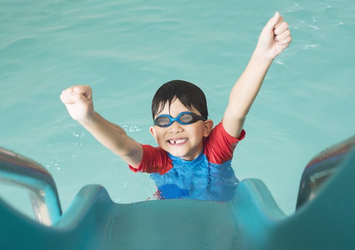 Os ensinamentos que o esporte traz para a vida das crianças; garoto na piscina com óculos de natação levanta os braços para o alto