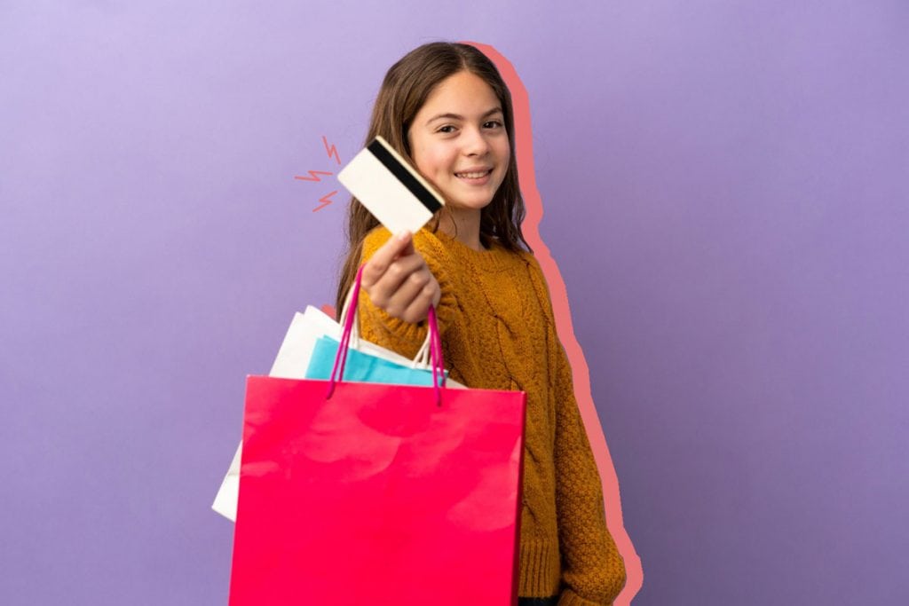 A importância da mesada para a educação financeira das crianças; menina sorrindo segurando um cartão de crédito preto e branco com duas sacolas penduradas no braço, uma azul e outra rosa