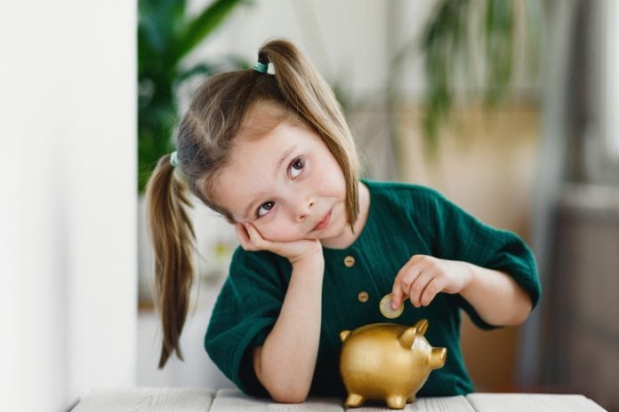 Como definir o valor da mesada das crianças?; Menina de cabelo preso com 'pitós' apoia bocheca sobre mão e coloca moeda em cofrinho
