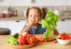 Dieta plant-based é alternativa saudável para alimentação das crianças; Menina de blusa azul segurando uma alface em uma das mãos, sentada à mesa com um recipiente com vegetais no canto esquerdo da imagem.