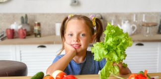 Dieta plant-based é alternativa saudável para alimentação das crianças; Menina de blusa azul segurando uma alface em uma das mãos, sentada à mesa com um recipiente com vegetais no canto esquerdo da imagem.