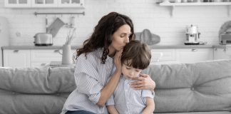 Dia de Finados: como retratar a morte com as crianças?; mãe abraçando filho e beijando sua cabeça, criança está triste e olhando para baixo, ambos estão sentados em um sofá cinza