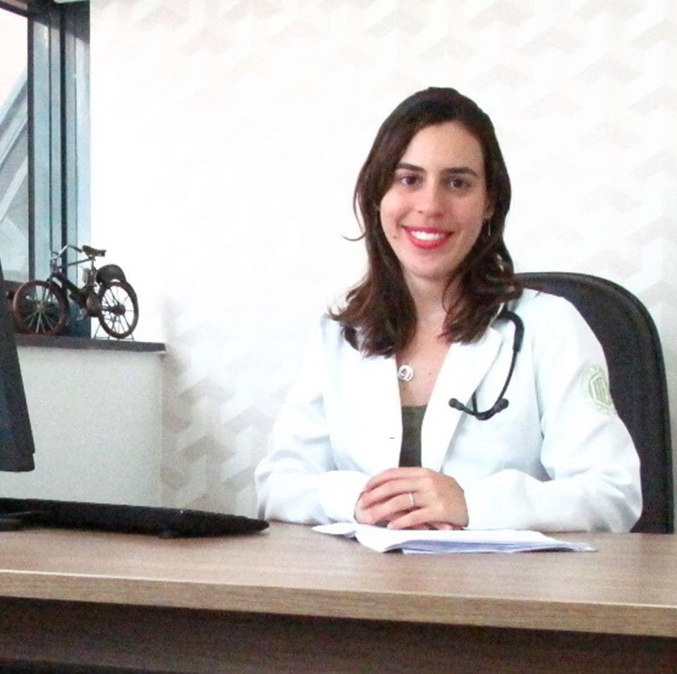 Infectologista Claudia Mello de jaleco branco, sentada em sua mesa.