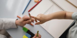 Mãos de criança seguram lápis e mão de adulto aponta para um dos lápis
