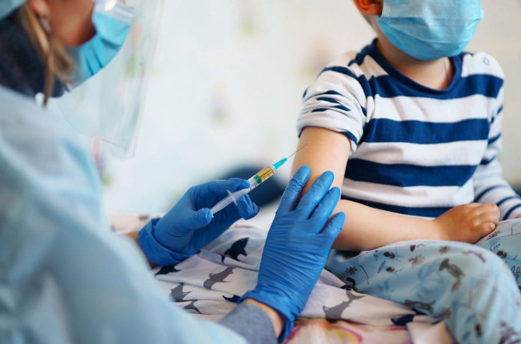 Vacinação contra o coronavírus: e a vez das crianças?; médica de luvas e máscara azuis aplicando vacina em criança de blusa azul listrada usando máscara