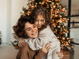 Mãe e filha se abraçam agachadas em frente à árvore de Natal