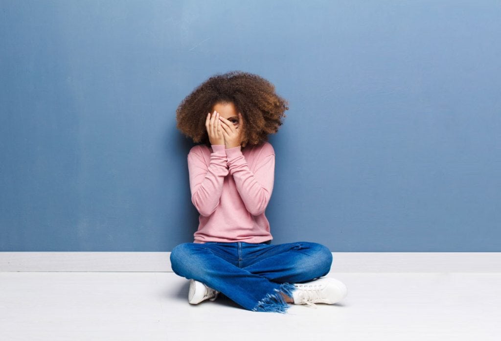 Introversão não é doença: as qualidades e desafios da criança introvertida; criança sentada de pernas cruzadas cobrindo o rosto com as mãos