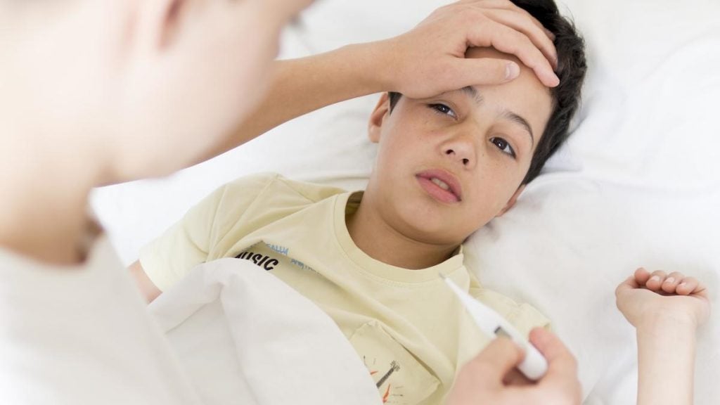 Adulto apoia mão na testa de menino e olha para termômetro na outra mão; Surto de gripe em SP: crescem casos em crianças