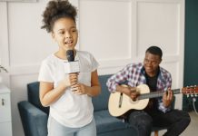 Menina de cabelo crespo canta, em pé, segurando microfone e pai no sofá toca violão