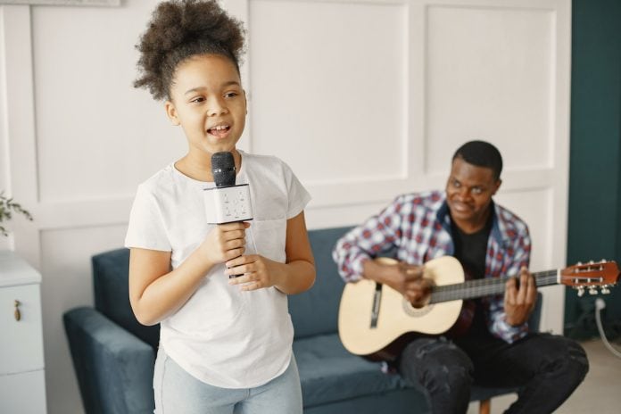 Menina de cabelo crespo canta, em pé, segurando microfone e pai no sofá toca violão