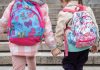 Duas garotas de costas, usando mochilas escolares, dando as mãos