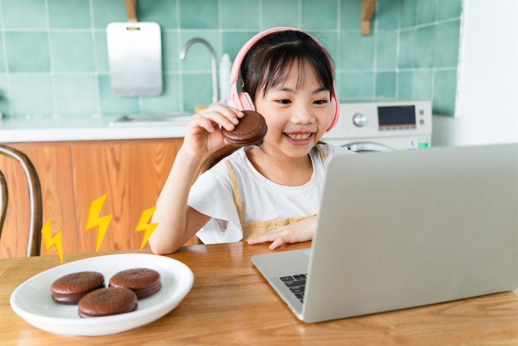 Menina asiática de cabelos lisos e fone de ouvido rosa comendo biscoitos de chocolate no café da manhã enquanto assiste às aulas em seu computador