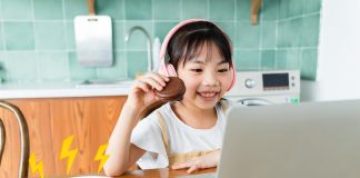 Menina asiática de cabelos lisos e fone de ouvido rosa comendo biscoitos de chocolate no café da manhã enquanto assiste às aulas em seu computador