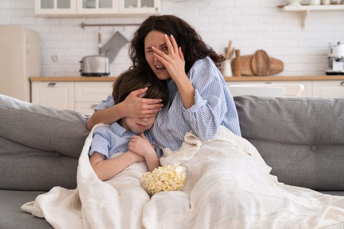 Mãe e filho sentados em um sofá, cobertos com uma manta, cobrindo os olhos, com feição de medo