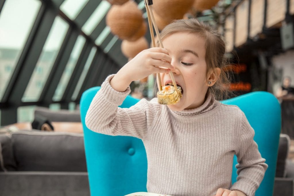 Criança sentada, com hashi na mão, comendo comida japonesa