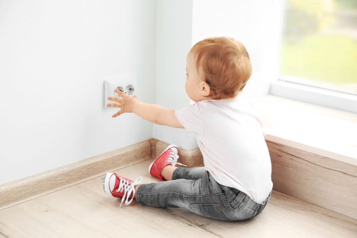 Criança sentada no chão estica braço em direção à tomada na parede