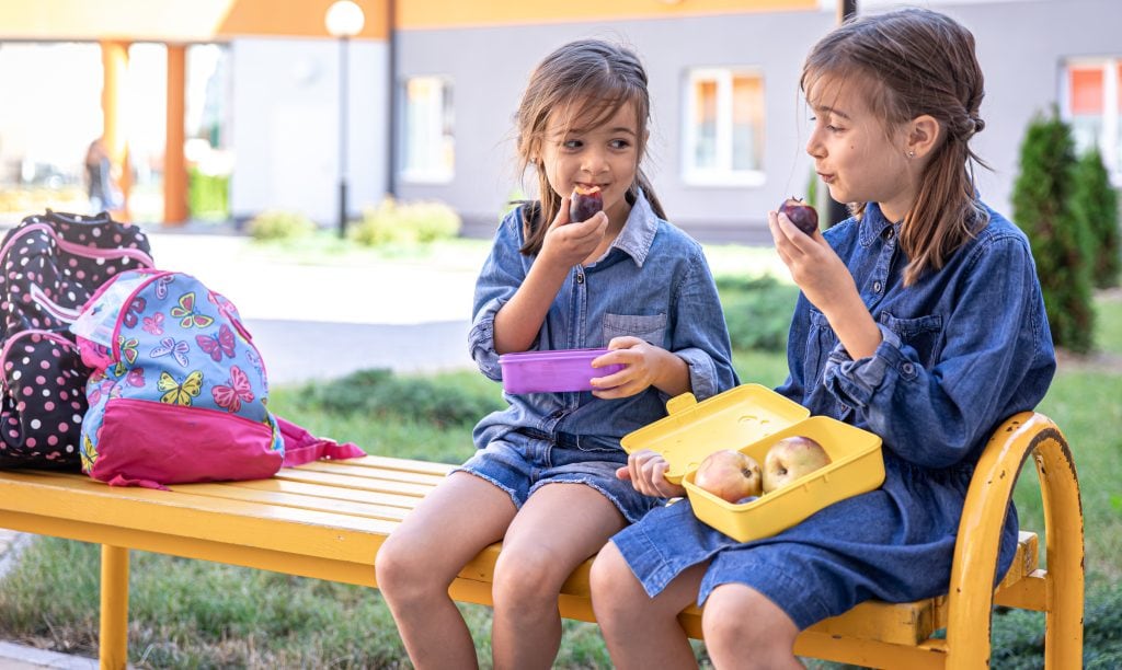 Duas meninas comem lanche em banco ao ar livre