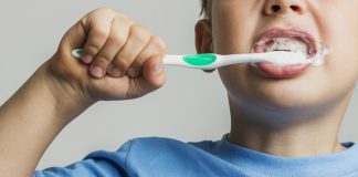 Menino de camiseta azul escova os dentesexcesso de creme dental, que pode provocar fluorose dentária nas crianças; imagem mostra rosto de menino escovando os dentes
