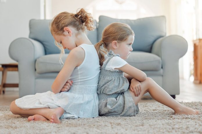 Briga entre irmãos: meninas estão sentadas no chão com as costas apoiadas uma na outra com cara de bravas