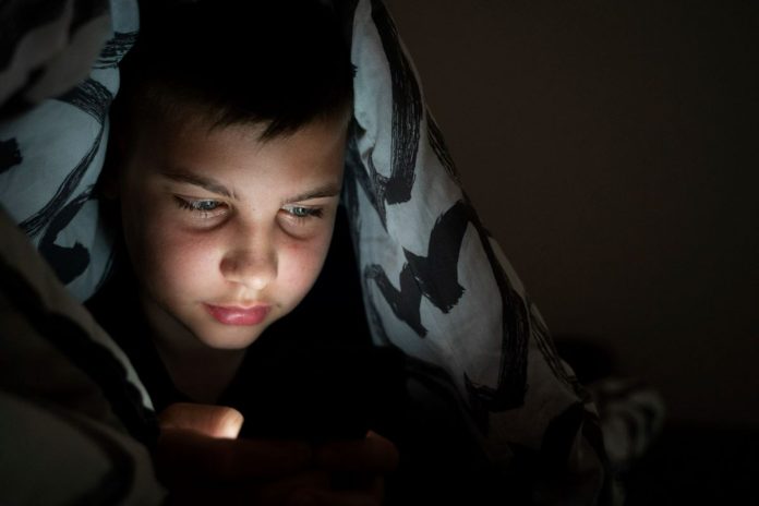 A geração do quarto vive plugada à internet, como esse menino, que está coberto por edredon, e olha tela de celular no escuro