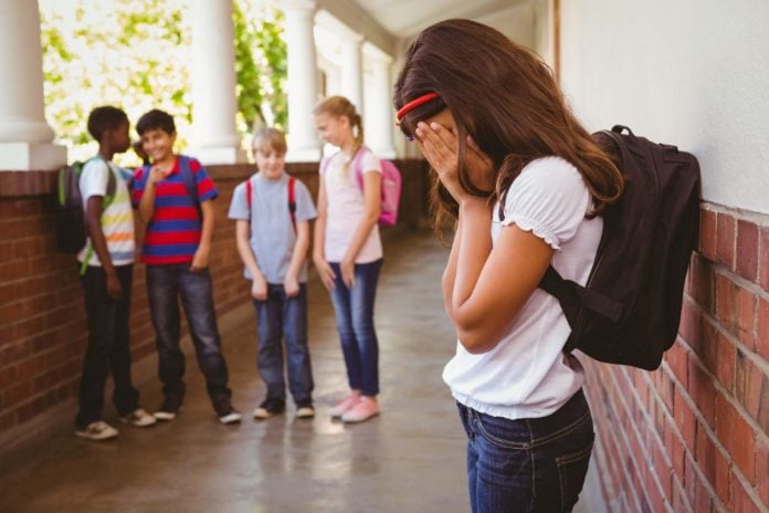 Menina de mochila nas costas tampa rosto com a mão e amigos olham para ela na escola