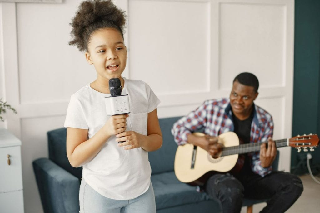 Menina canta no microfone e pai ao fundo toca violão