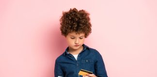 Menino de cabelos cacheados ruivos segura numa mão um cartão de crédito e na outra o celular