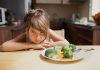 Menina encosta braços sobre mesa ao lado de prato com brócolis e legumes