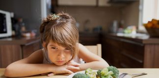 Menina encosta braços sobre mesa ao lado de prato com brócolis e legumes