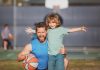Pai agachado com bola na mão posa para foto abraçando filho em quadra de esportes