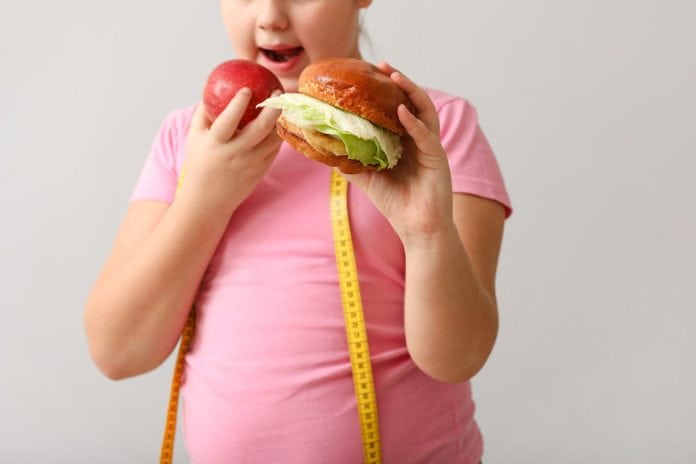 Menina gordinha tem fita métrica pendurada no ombro e segura uma maçã numa mão e hambúrguer em outra
