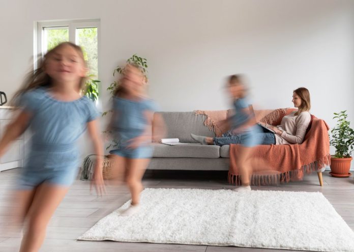 Criança corre ao redor do tapete da sala, mãe está no sofá