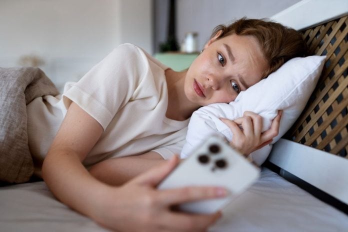 Menina deitada na cama olha para tela de celular que segura na mão