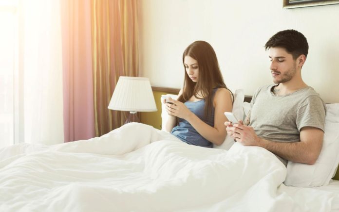 Casal sentado na cama, cada um olha para o seu celular