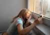 Menina adolescente encosta a cabeça junto à janela e olha para o celular
