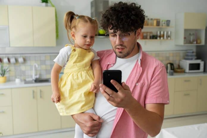 Pai segura filhos nos braços e olha para tela de celular