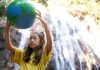 Menina segura globo terrestre com as mãos ao alto em frente a cachoeira