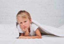 Criança com cara chateada em cima da cama embaixo do corbertor; Sono infantil: como fazer a criança dormir