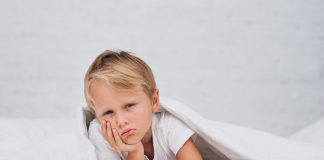 Criança com cara chateada em cima da cama embaixo do corbertor; Sono infantil: como fazer a criança dormir
