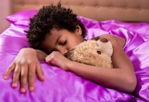 Menina afro dorme abraçada a ursinho em cama com lençol lilás