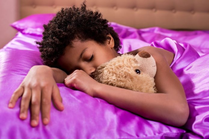 Menina afro dorme abraçada a ursinho em cama com lençol lilás