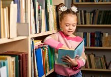 Menina lê livro em pé, apoiada em estante de biblioteca
