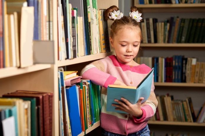 Menina lê livro em pé, apoiada em estante de biblioteca